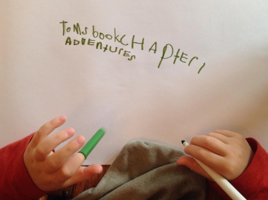 Image shows a child's hand holding a pen. On a piece of paper is written 'Tom's book Chapter 1 adventures'