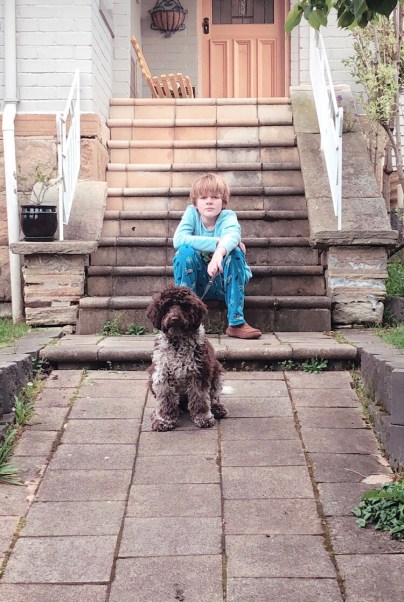 A boy sitting on the front steps of a house, holding the lead of a brown and white poodle puppy