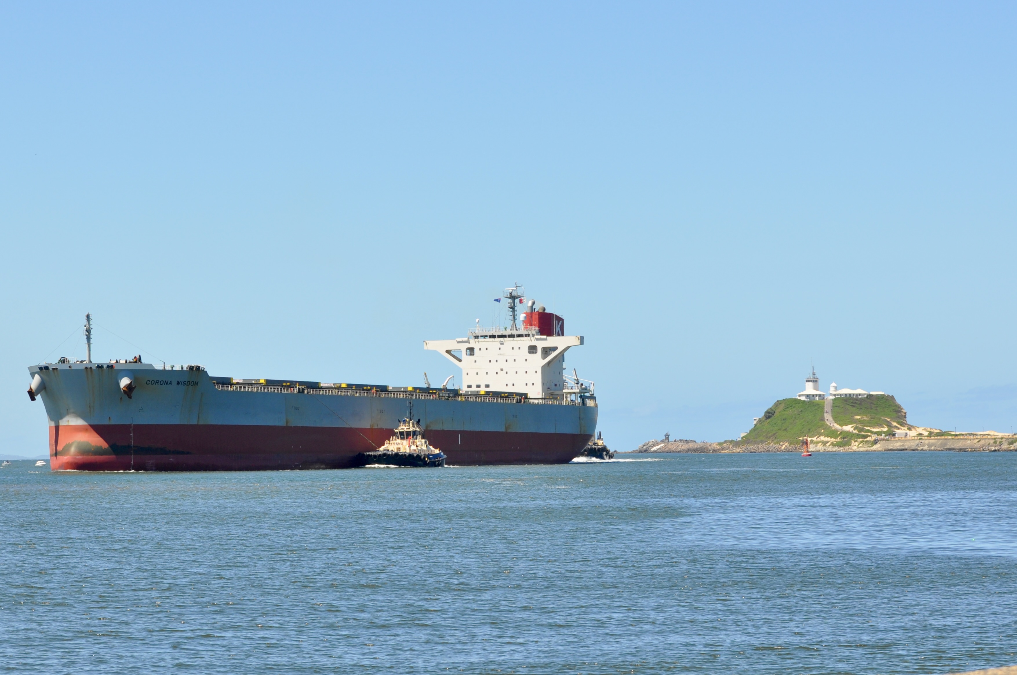 Photo shows a bulk carrier accompanied by a tug coming into the port of Newcastle in New South Wales. Nobby's Lighthouse is visible in the background.