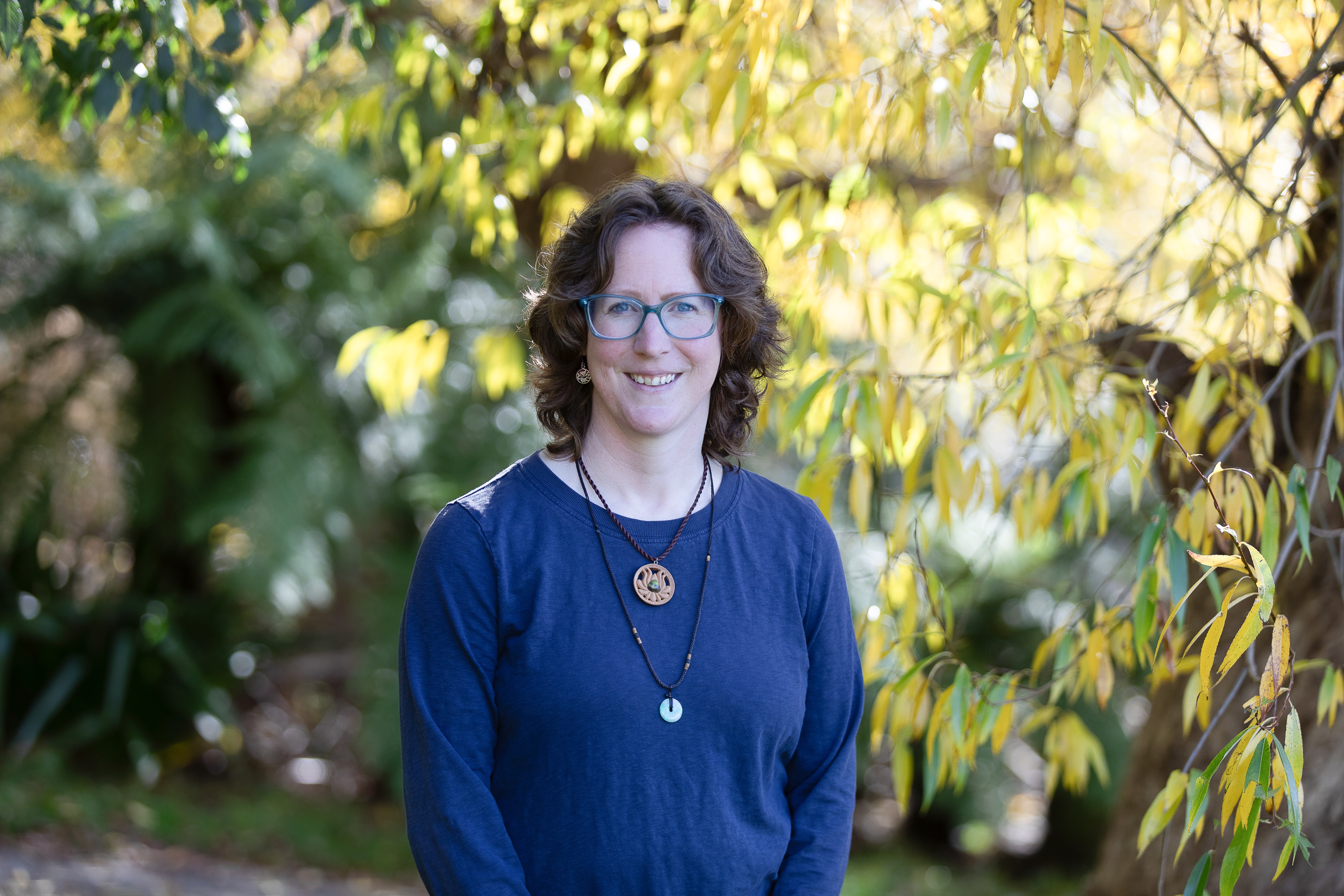 Ruth Dawkins DorkyMum - a white woman with brown hair, wearing a blue top and a wooden necklace, stands in front of some pretty trees
